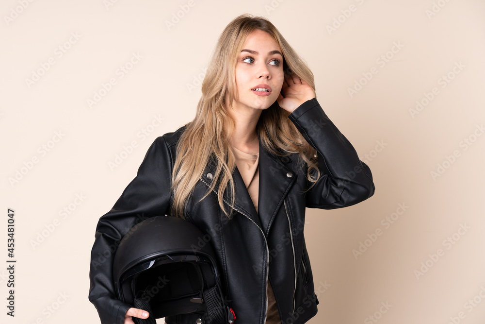 Russian girl with a motorcycle helmet isolated on beige background listening to something by putting hand on the ear Russian girl with a motorcycle helmet isolated on beige background listening to something by putting hand on the ear