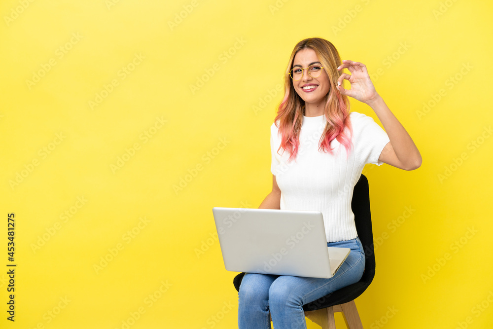 Naklejka premium Young woman sitting on a chair with laptop over isolated yellow background showing ok sign with fingers
