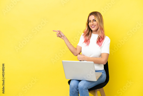 Young woman sitting on a chair with laptop over isolated yellow background pointing finger to the side