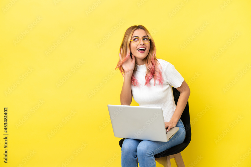 Young woman sitting on a chair with laptop over isolated yellow background listening to something by putting hand on the ear Young woman sitting on a chair with laptop over isolated yellow background listening to something by putting hand on the ear