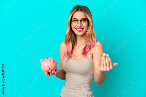 Young woman over isolated blue background holding a piggybank inviting to come with hand. Happy that you came