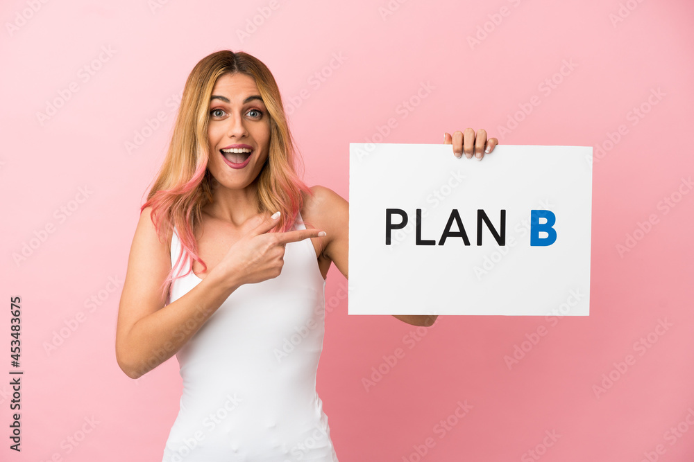 Young woman over isolated pink background holding a placard with the message PLAN B with surprised expression Young woman over isolated pink background holding a placard with the message PLAN B with surprised expression
