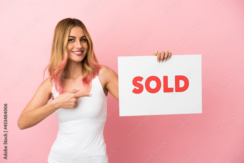Young woman over isolated pink background holding a placard with text SOLD and pointing it Young woman over isolated pink background holding a placard with text SOLD and pointing it