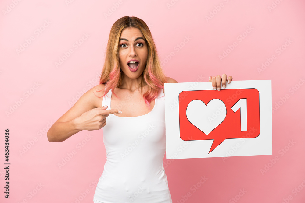 Young woman over isolated pink background holding a placard with Like icon and pointing it Young woman over isolated pink background holding a placard with Like icon and pointing it