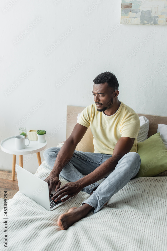 barefoot african american freelancer typing on laptop in bedroom
