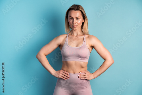 Bodybuilding and sport concept. Young fit pretty woman with short hair dressed in sport clothes putting her arms into the waist and posing against blue blank wall © speed300