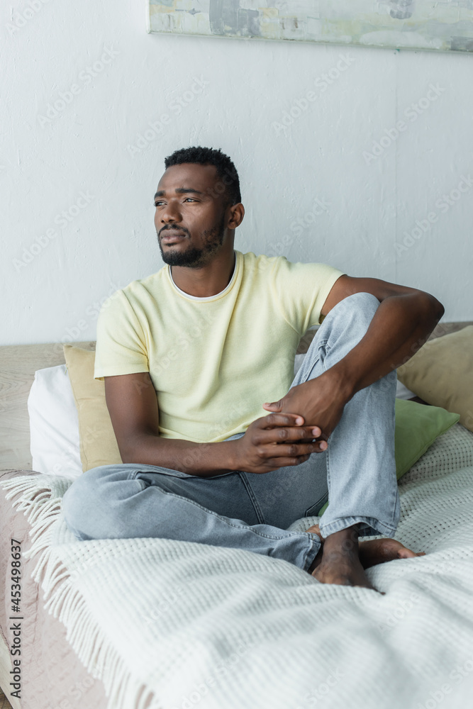 barefoot african american man looking away while thinking and sitting on bed