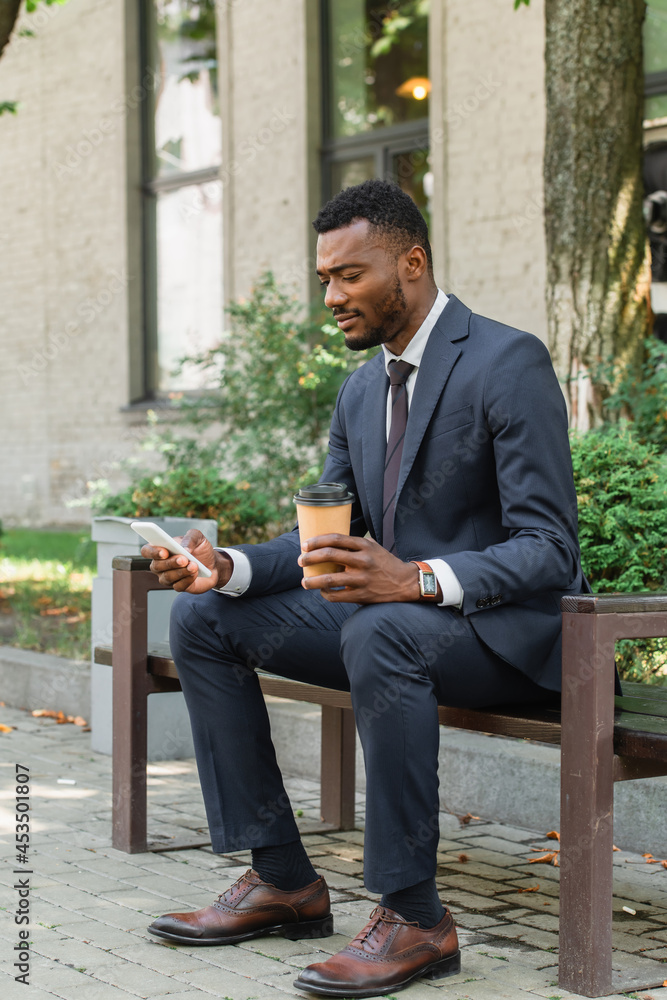 Obraz premium african american businessman using mobile phone and holding coffee to go and sitting on bench