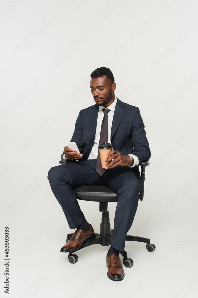african american businessman in suit sitting in office chair, messaging on smartphone and holding paper cup isolated on grey african american businessman in suit sitting in office chair, messaging on smartphone and holding paper cup isolated on grey