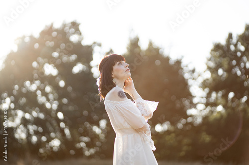 Outdoor summer shot of native american indian woman, wearing white boho dress, feather accessories in hair and tattoos on her body, looking up. Trees background with free text space