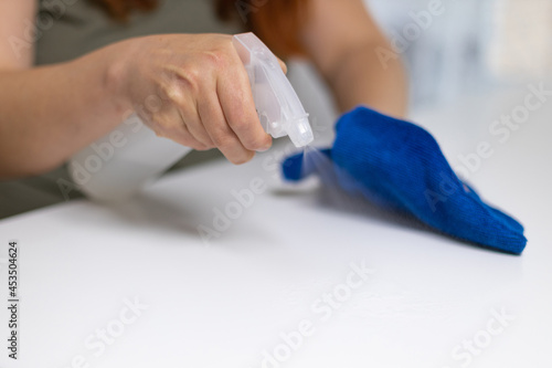 Young woman housekeeper is cleaning, wiping down table surface with blue gloves, wet rag, spraying bottle cleaner. COVID-19 prevention sanitizing inside