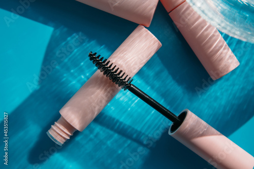 pink clean makeup brush for mascara lies on the open tube, near to closed tubes of cosmetics, lip gloss, liquid lipstick, pink eyeliner, glass vase on a blue background with shadows. Copy space