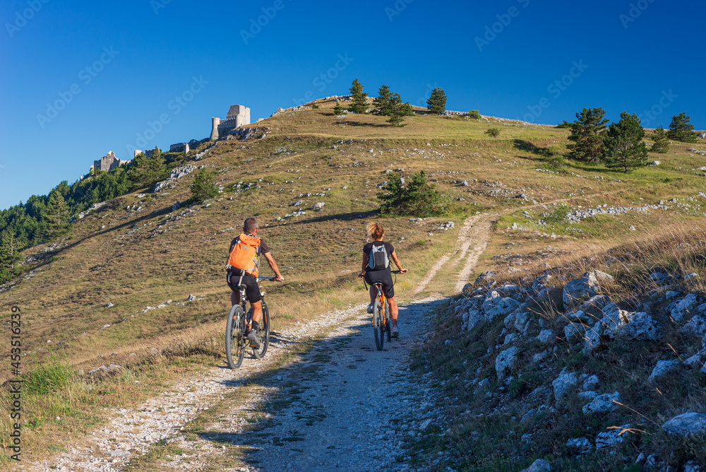 Obraz premium Man and woman riding mtb to castle ruins on mountain top at Rocca Calascio, italian travel destination, Gran Sasso National Park, Abruzzo, Italy. Clear blue sky