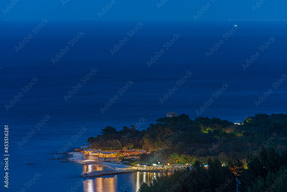 Portonovo village view from above by night. Illuminated restaurants and beach resort reflection on blue sea. Italy Marche tourism destination