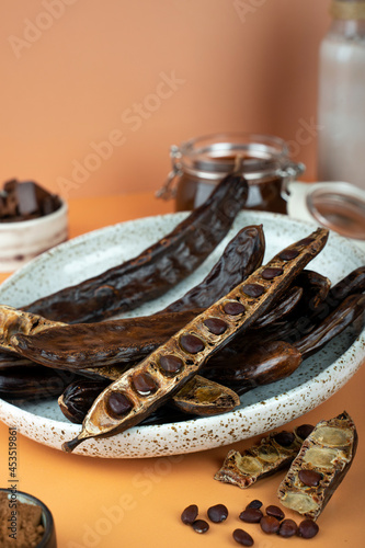 Organic carob pods, seeds in a ceramic plate on a beige background, locust bean healthy food, Ceratonia siliqua harnup. Natural vegan eating. creative food background. Copy space. Top view photo