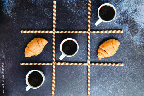 Businessman working morning with cups of hot coffee and sweet croissants on black background. Game of noughts and crosses. Top view, copy space. Flat lay. Creative concept. Tic tac toe photo