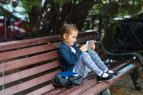a little girl in a school uniform  sitting on a bench  and holding a tablet in her hands