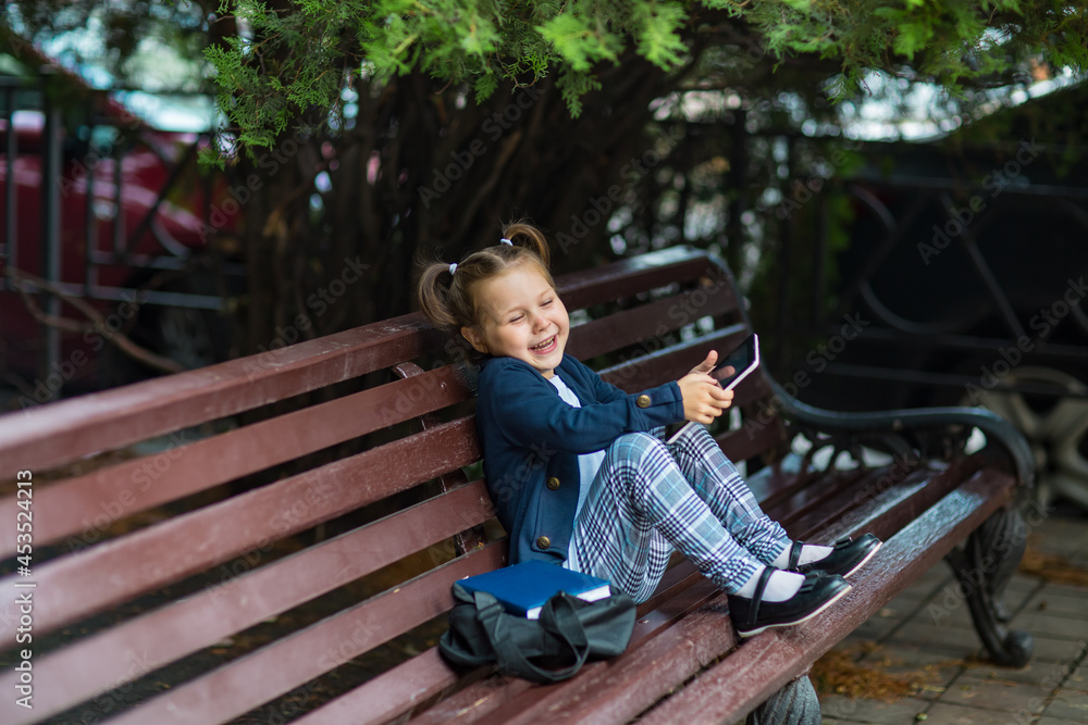 a little girl in a school uniform, sitting on a bench, and holding a tablet in her hands