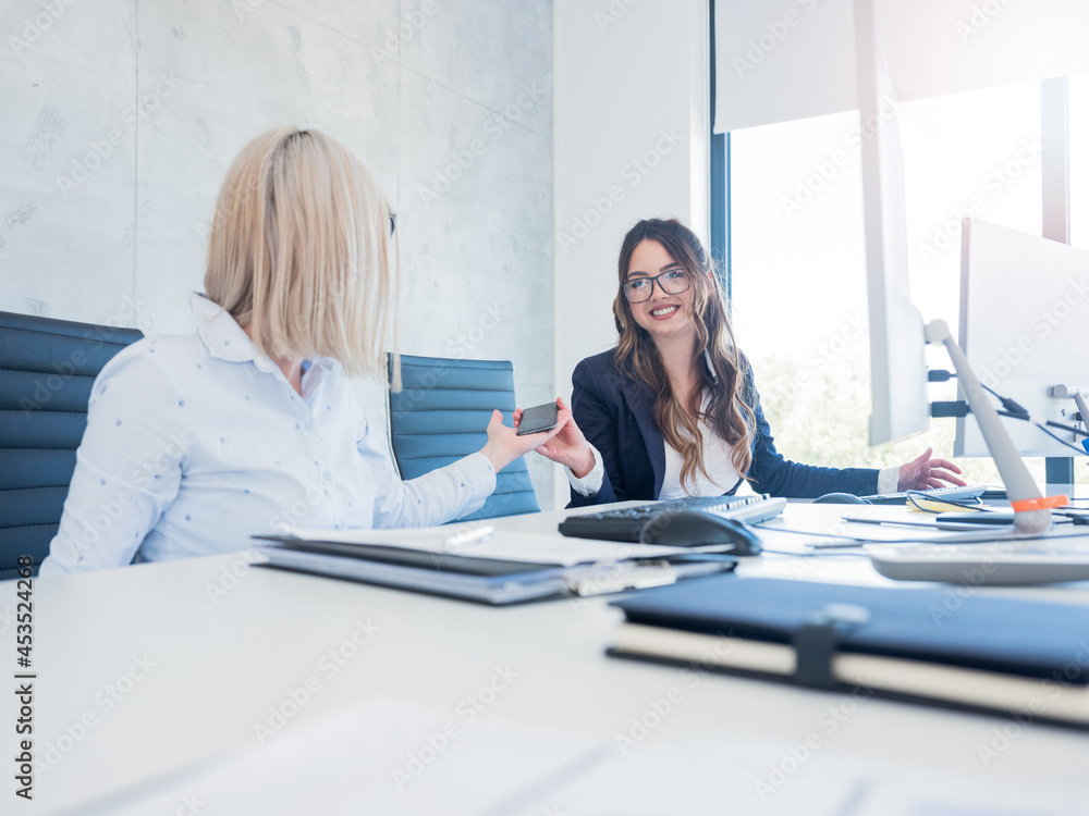 Young brunette giving her colleague a phone, casual conversation between coworkers in office. Young brunette giving her colleague a phone, casual conversation between coworkers in office.