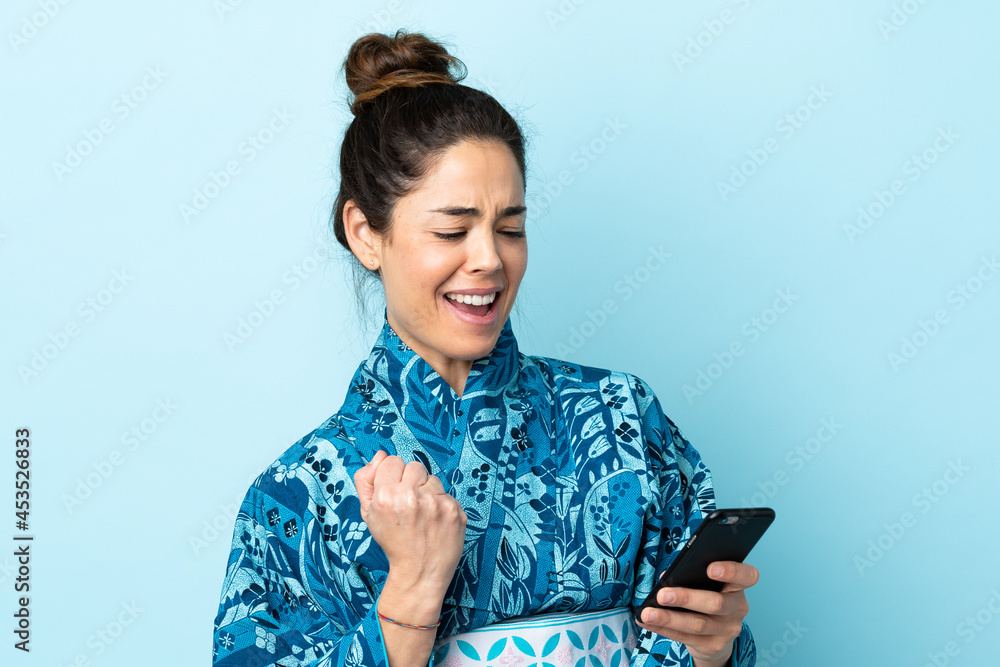 Woman wearing kimono over isolated background with phone in victory position Woman wearing kimono over isolated background with phone in victory position