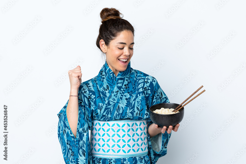 Woman wearing kimono over isolated white background celebrating a victory while holding a bowl of noodles with chopsticks Woman wearing kimono over isolated white background celebrating a victory while holding a bowl of noodles with chopsticks