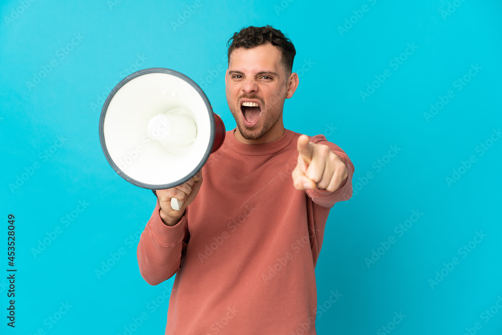 Young caucasian handsome man isolated on blue background shouting through a megaphone to announce something while pointing to the front Young caucasian handsome man isolated on blue background shouting through a megaphone to announce something while pointing to the front