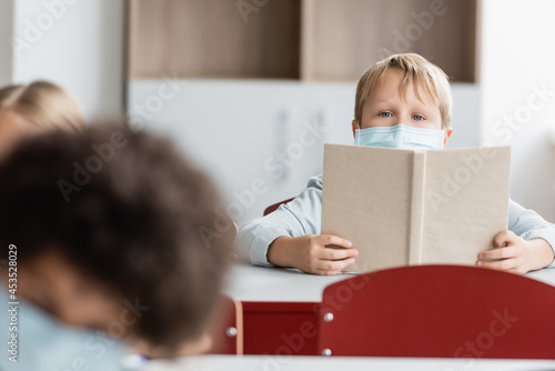 schoolchild in medical mask holding book while looking at camera during lesson