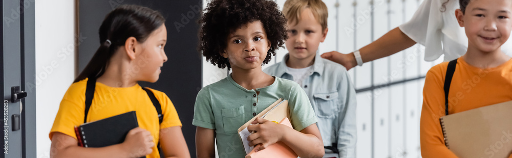 pensive african american schoolboy holding notebooks near multiethnic pupils and teacher, banner pensive african american schoolboy holding notebooks near multiethnic pupils and teacher, banner