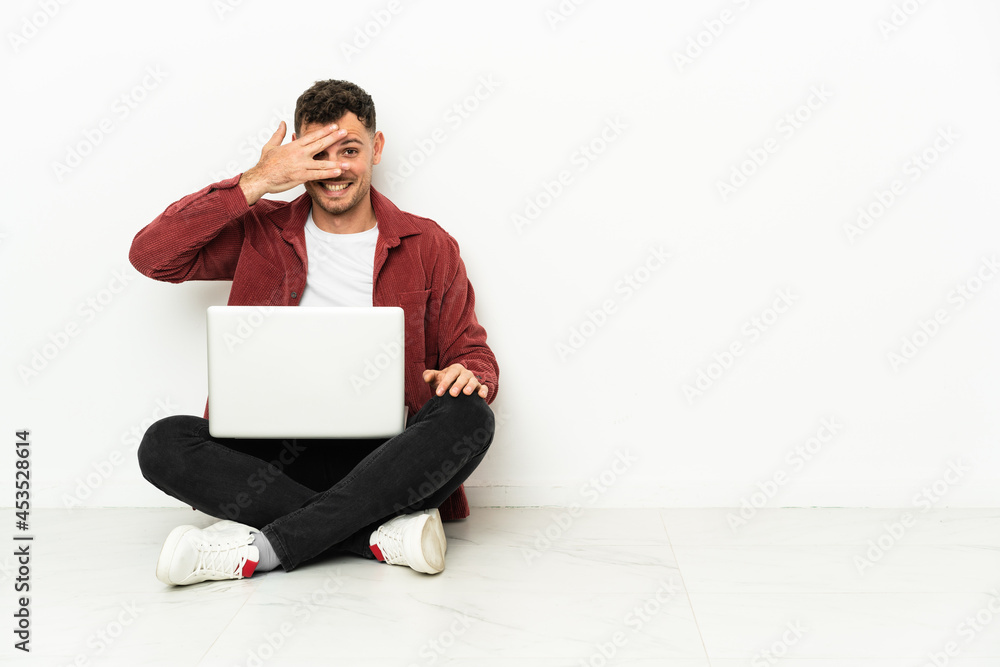 Young handsome caucasian man sit-in on the floor with laptop covering eyes by hands and smiling Young handsome caucasian man sit-in on the floor with laptop covering eyes by hands and smiling