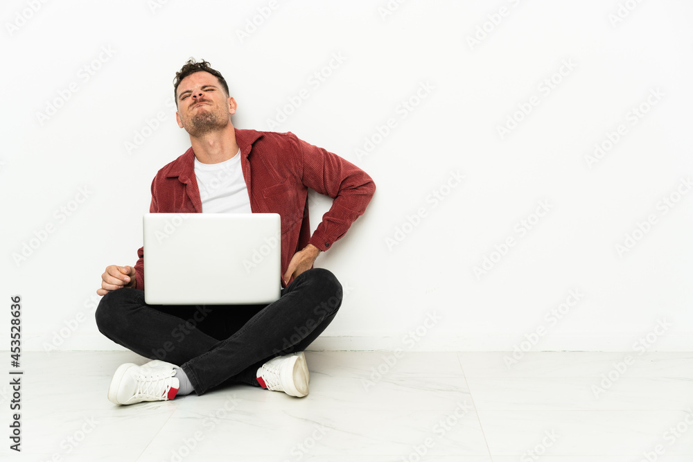 Young handsome caucasian man sit-in on the floor with laptop suffering from backache for having made an effort Young handsome caucasian man sit-in on the floor with laptop suffering from backache for having made an effort