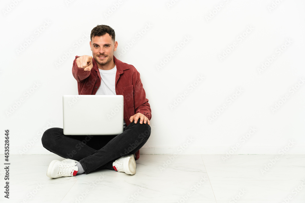 Young handsome caucasian man sit-in on the floor with laptop points finger at you with a confident expression Young handsome caucasian man sit-in on the floor with laptop points finger at you with a confident expression
