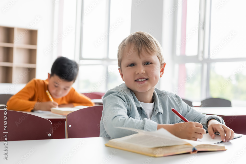 happy schoolkid smiling at camera near asian boy writing on blurred background happy schoolkid smiling at camera near asian boy writing on blurred background