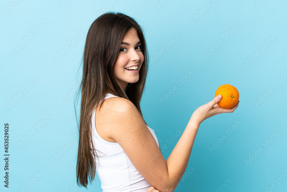 Teenager Brazilian girl over isolated blue background holding an orange Teenager Brazilian girl over isolated blue background holding an orange