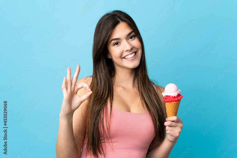 Teenager Brazilian girl holding a cornet ice cream over isolated blue background showing ok sign with fingers Teenager Brazilian girl holding a cornet ice cream over isolated blue background showing ok sign with fingers