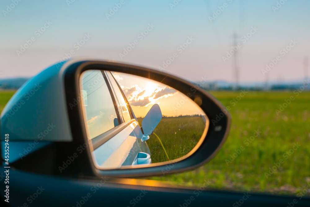 Rear mirror with a reflection of the female silhouette charging an electric car, in the background visible sky at sunset