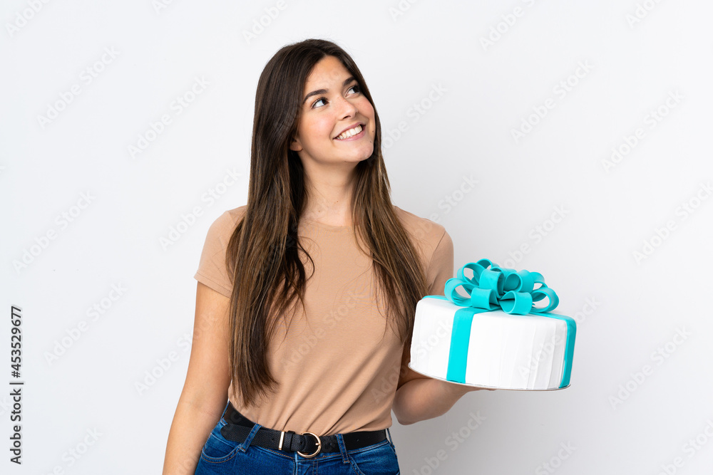 Teenager pastry chef holding a big cake over isolated white background looking up while smiling Teenager pastry chef holding a big cake over isolated white background looking up while smiling