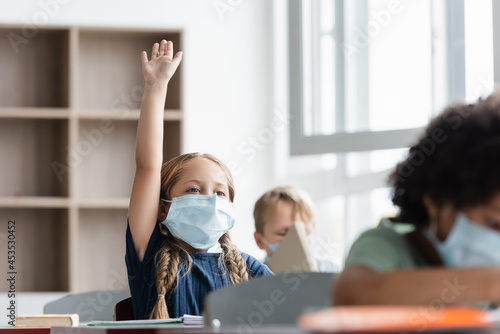 schoolgirl in medical mask raising hand during lesson near multicultural classmates