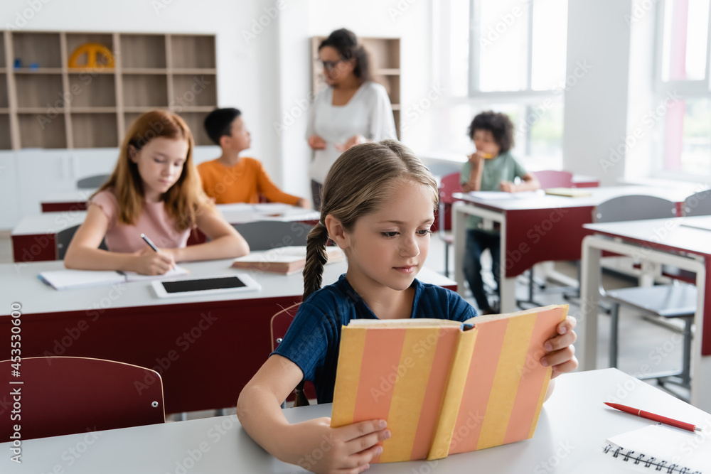 Obraz premium girl reading book near multiethnic classmates and african american teacher on blurred background
