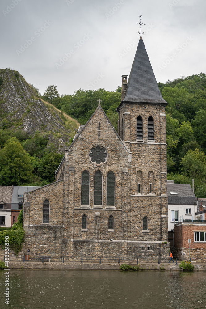 Dinant, Wallonia, Belgium - August 8, 2021: Church of Saint Paul des Rivages along Meuse river with green forested hill and steep descending rock formations in back.