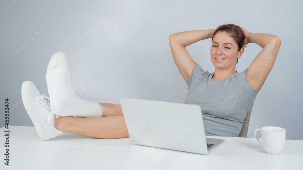 Caucasian woman lifted her leg with plaster to work desk and works on laptop on white background Caucasian woman lifted her leg with plaster to work desk and works on laptop on white background