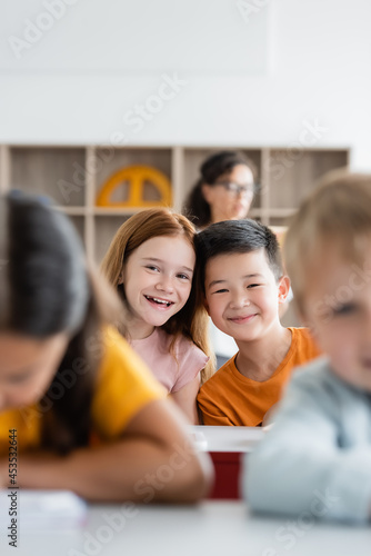 cheerful interracial kids smiling at camera near blurred classmates in classroom