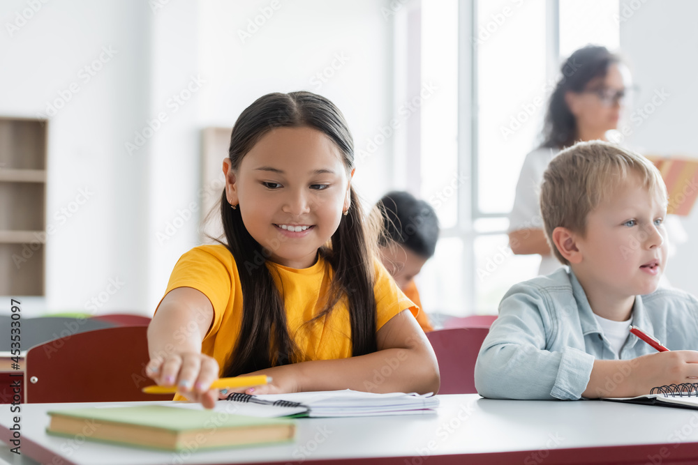 cheerful asian schoolgirl reaching book while sitting at desk near classmate cheerful asian schoolgirl reaching book while sitting at desk near classmate