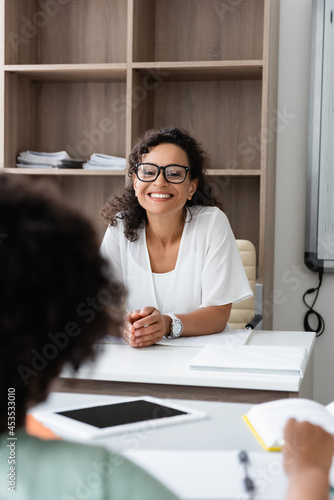 cheerful teacher looking at african american boy on blurred background in classroom