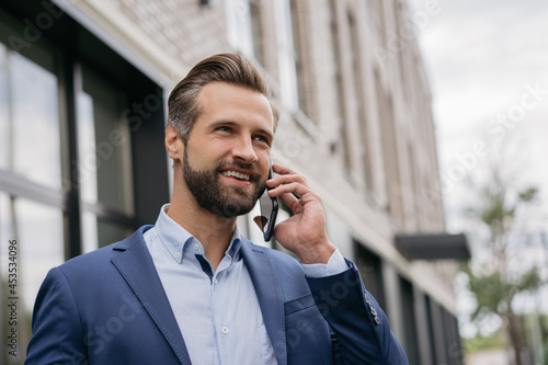 Portrait of handsome businessman wearing stylish suit talking on mobile phone, standing on the street looking at copy space. Successful business 