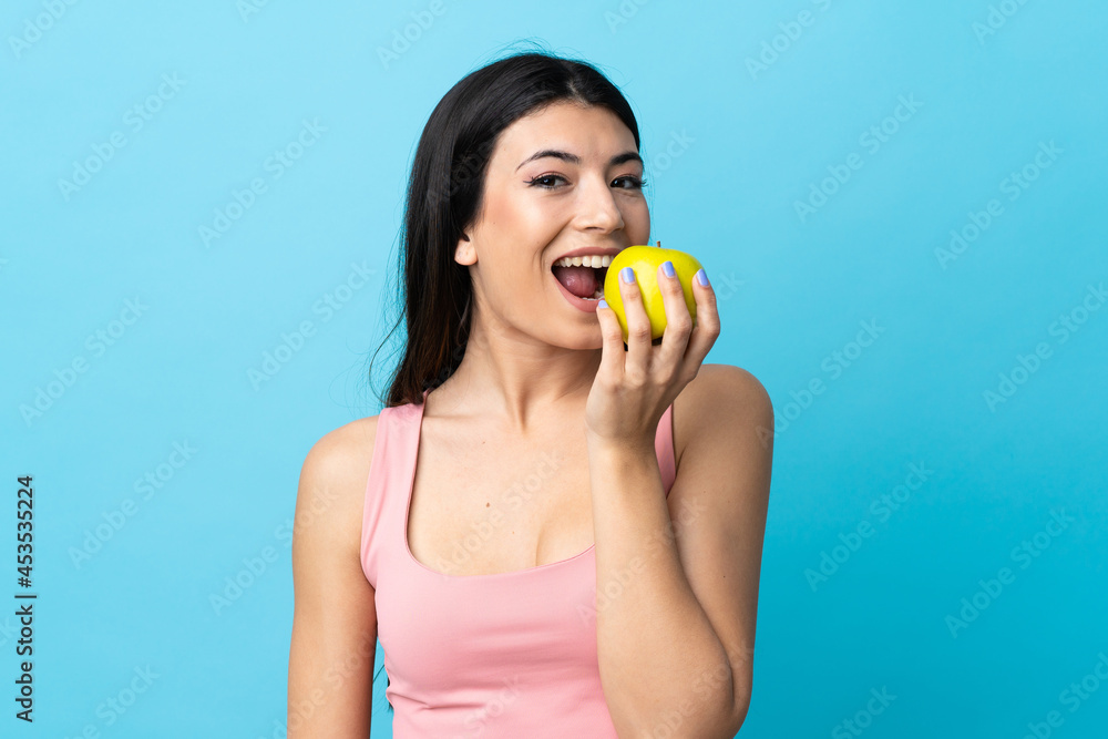 Young brunette girl over isolated blue background eating an apple Young brunette girl over isolated blue background eating an apple