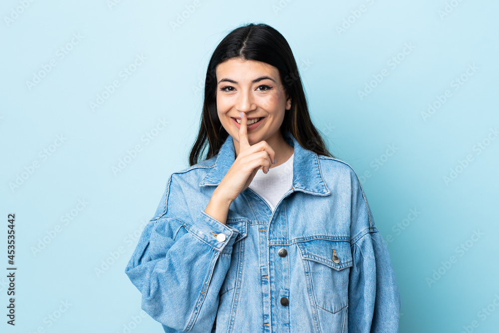 Young brunette girl over isolated blue background showing a sign of silence gesture putting finger in mouth Young brunette girl over isolated blue background showing a sign of silence gesture putting finger in mouth