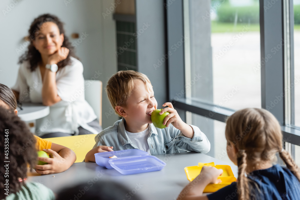 boy eating apple near classmates and blurred african american teacher boy eating apple near classmates and blurred african american teacher