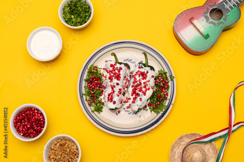 Flat lay photography with chiles en Nogada, a traditional dish from Puebla with the addition of walnut cream, pomegranate seeds and parsley, ingredients, toy guitar and little hat on yellow background photo