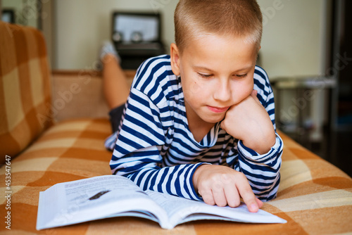 Cute ten year old boy doing homework at home on couch lying on his back during pandemic or after school. Selective focus. Close-up. Portrait of caucasian schoolboy boy. Spends time usefully at leisure