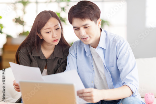 Young husband and wife holding paperwork and using laptop at home.